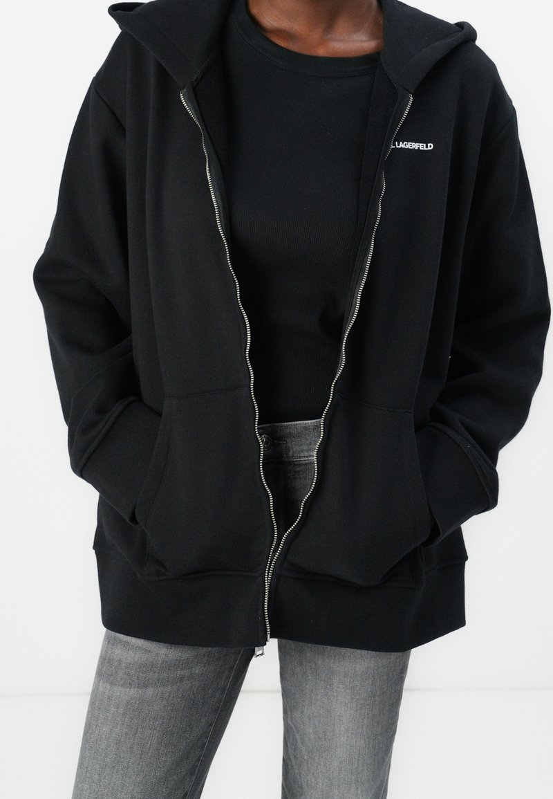 Personne portant un sweat à capuche zippé noir avec les mains dans les poches, par-dessus un t-shirt noir, associé à un jean gris en denim.