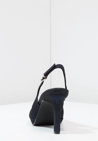 Zapato de tacón negro con parte superior de tela texturizada que presenta manchas azules, con la parte trasera abierta, correa ajustable y un tacón stileto delgado.