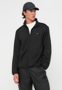 Black zip-up fleece jacket with high collar, smooth texture, and small logo detail. Paired with a dark cap and matching trousers.