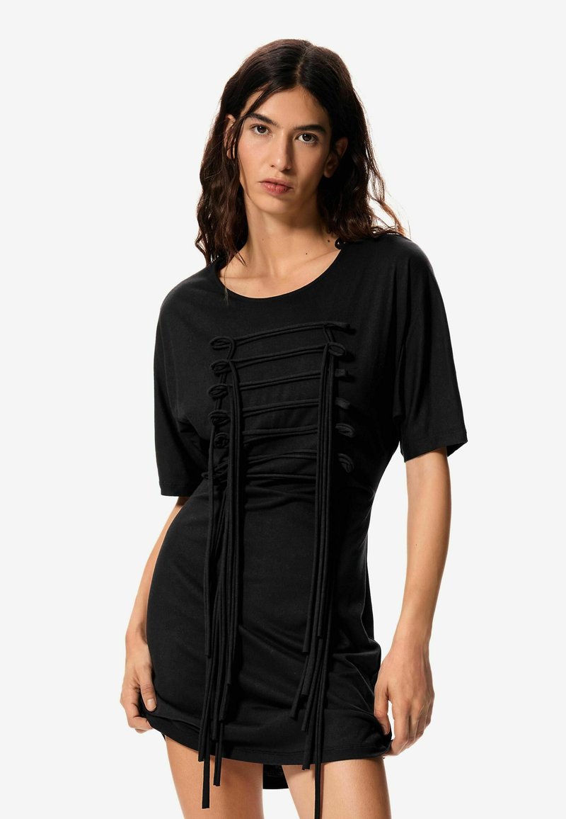 Femme aux longs cheveux noirs portant une robe courte noire avec des détails de cordon noué sur le devant, debout devant un fond blanc.