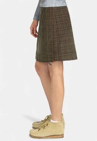 Persoon in een olijfgroen geruit knielang rokje, beige enkellaarsjes, en een blauw-wit gestreept shirt met lange mouwen.