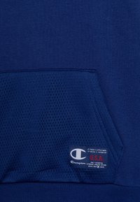 Tessuto blu con una tasca texturizzata che presenta il logo Champion e il testo rosso "U.S.A." su una piccola etichetta cucita.