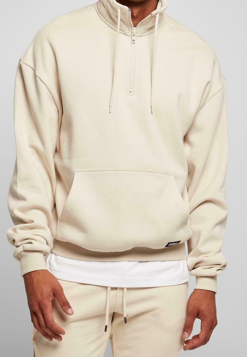 Uomo che indossa un maglione beige a zip con tasca frontale e pantaloni beige abbinati, con una maglietta bianca visibile sotto l'orlo del maglione.