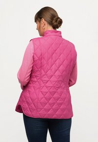 Prošívaná růžová vesta s vysokým límcem a diamantovým vzorem, vrstvená přes růžovou košili s dlouhým rukávem. Na spodní části jsou vidět džíny.