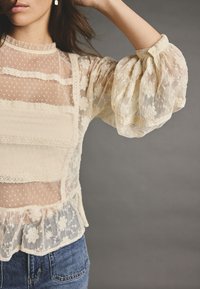Blusa in crema trasparente con motivi floreali in pizzo, inserti a pois e maniche a sbuffo; orlo arricciato e polsini con bottoni. Jeans in denim visibili sotto.