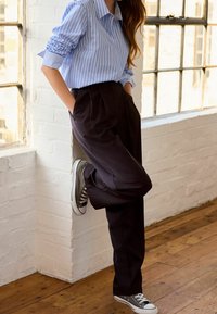 Chemise à rayures bleues et blanches à boutons avec manches retroussées, associée à un pantalon noir plissé et des baskets Converse grises.