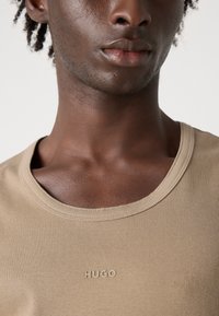 Tricou ribbed bej cu guler rotund și logo-ul „HUGO” embosat pe față, fabricat din material de bumbac moale.