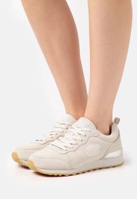 Beige sneakers made of suede with a textured surface. Features white laces, silver accents, and a rubber sole with a gum-colored tread.
