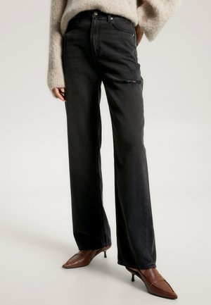 Personne portant un jean noir taille haute à jambes larges avec une légère usure subtile et des chaussures marron à talons kitten pointues, debout avec une main dans la poche.