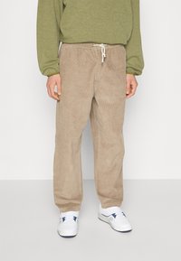 Beige kordflätade byxor med avslappnad passform, med en elastisk midja och dragsko. Matchas med en grön sweatshirt och vita sneakers.