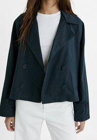 Granatowa, dwurzędowa kurtka z szerokim kołnierzem, z guzikami i skróconym krojem, w połączeniu z białą bluzką i jasnymi dżinsami.