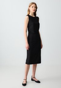 Schwarzes, ärmelloses, knielanges Kleid mit vertikalen goldenen Streifen; glattes Material; kombiniert mit schwarzen Schuhen mit Knöchelriemen.