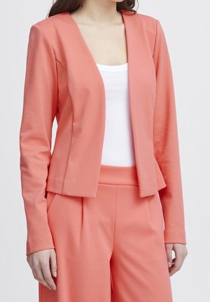Vrouw draagt een zalmroze blazer en bijpassende broek over een wit topje met ronde hals, staand tegen een effen achtergrond.