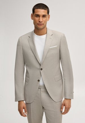 Man draagt een licht beige colbert en bijpassende broek over een wit shirt met ronde hals, staand tegen een effen achtergrond.