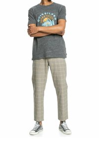 Persona con una camiseta gráfica gris de Quiksilver, pantalones de cuadros beige y zapatillas bajas negras, de pie con los brazos cruzados.