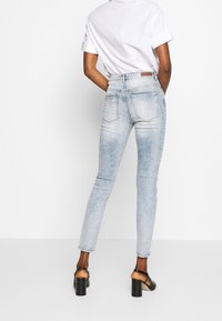 Jeans de tiro alto color azul claro con una textura desgastada, ajuste slim y dos bolsillos traseros. Combinados con una camiseta blanca y sandalias negras de tacón.