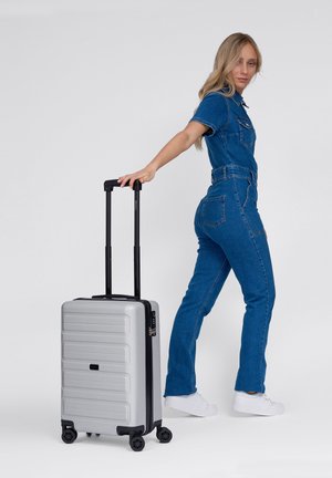Frau in einem Jeans-Jumpsuit, die einen silbernen Hartschalenkoffer mit vier Rollen und einem ausziehbaren Griff vor einem schlichten Hintergrund zieht.
