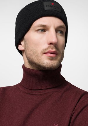 Czarna dzianinowa czapka beanie z prostokątną naszywką z logo, noszona z maroonowym swetrem z golfem, podkreślająca prążkowaną teksturę i osłonę szyi.