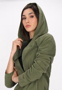 Oliwkowa bluza z kapturem na zamek, wykonana z miękkiego polaru, z kapturem i dopasowanymi rękawami, noszona na czarnej bluzce, model trzymający materiał.