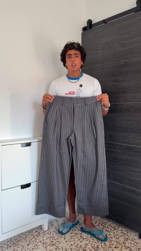 Grijze krijtstreepjasje met een geplooide voorkant, brede benen en een tailleband. Vastgehouden door een persoon die een wit shirt en slippers draagt.