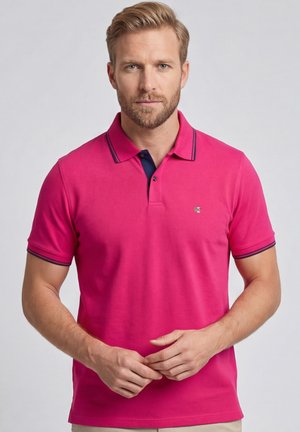 Blonde man met baard die een felroze poloshirt draagt met marineblauwe bies bij de kraag en mouwen, staand tegen een effen achtergrond.