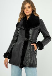 Cappotto in pelle nera con ampio colletto e polsini in pelliccia, vita cinturata e dettagli a coste sulle maniche, dotato di tasche e una superficie liscia.