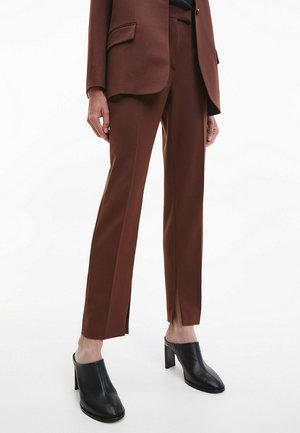 Frau trägt braune, maßgeschneiderte Hose mit Frontal-Schlitzen, kombiniert mit einem passenden Blazer und schwarzen, hochhackigen Slip-On-Schuhen vor weißem Hintergrund.