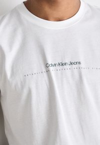 Camiseta de algodón blanca con el logo "Calvin Klein Jeans" en verde, con el texto "ESTABLECIDO EN MIL NOVECIENTOS SETENTA Y OCHO" abajo. Textura suave.