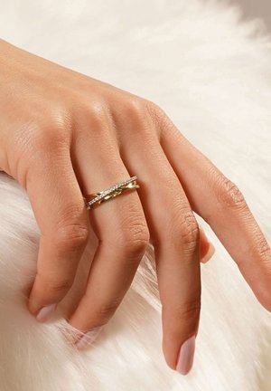 Goldfarbener Ring mit verschlungenen Bändern, von denen eines mit kleinen klaren Edelsteinen besetzt ist, auf einer Hand, die vor einem weichen weißen Fellhintergrund ruht.