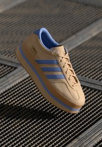 Tan nahast tossud siniste aktsentidega, millel on kolm triipu, ümmargune varvas ja tekstureeritud tald. Bränding on kuvatud küljel ja keelel.