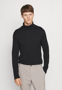 Camisa de manga larga de cuello alto negra con un diseño ajustado, textura suave y costuras mínimas, combinada con pantalones de color beige claro.