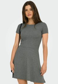 Femme avec de longs cheveux bruns portant une robe en tricot côtelé grise à manches courtes et portant un sac à épaule noir sur fond blanc.