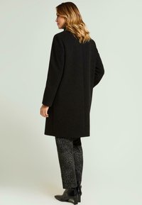 Abrigo negro largo con una textura suave y un corte entallado, combinado con pantalones oscuros estampados y botines. Diseño sencillo y elegante.