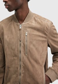 Giacca bomber in suede beige con una texture morbida, chiusura frontale con cerniera e tasca laterale con zip. Collo e polsini a costine per una vestibilità aderente.