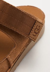 Brune semsket sandaler med ribbet tekstilrem og gummisåle. Har preget merkevarelogo på remmen og glatt innerside.