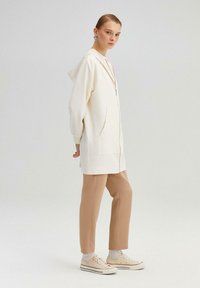 Jachetă albă cu glugă, cu buzunare frontale, închidere cu fermoar; asociată cu pantaloni bej și încălțăminte de culoare deschisă. Textură de material neted.