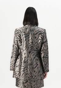 Person med rett, mørkt hår iført en metallisk blazer og skjørt sett med svart og bronse abstrakte zebra-striper, vender bort.
