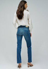 Jean en denim bleu avec une coupe slim et une légère évasement, associé à un top blanc texturé. La tenue comprend une étiquette beige et des talons à lanières.
