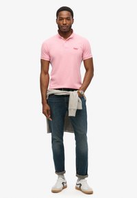 Różowa koszulka polo z małym logo, jasnoszare sportowe sweter związany w pasie, niebieskie dżinsy oraz białe sneakersy z szarymi i beżowymi akcentami.