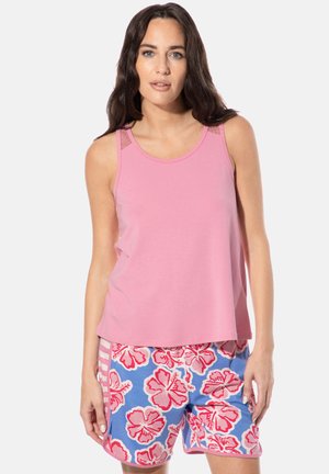 Frau trägt ein ärmelloses pinkes Oberteil und blaue Shorts mit pinken und roten Hibiskusblumenmuster, steht mit entspannten Armen.