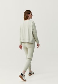 Ljusgrön sweatshirt och matchande åtsittande byxor i mjukt material. Modellen bär vita skor med brun sula. Enkelt, avslappnat design.