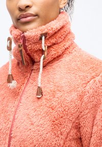 Γυναίκα που φοράει κοραλλί χνουδωτό μπουφάν με ψηλό γιακά, λευκά κορδόνια και φερμουάρ μπροστά, εμφανιζόμενη από τους ώμους μέχρι το πιγούνι.