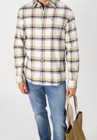 Béžová a námořnická kostkovaná košile s dlouhými rukávy, spárovaná s světle modrými džíny. V ruce je držena hnědá plátěná taška.