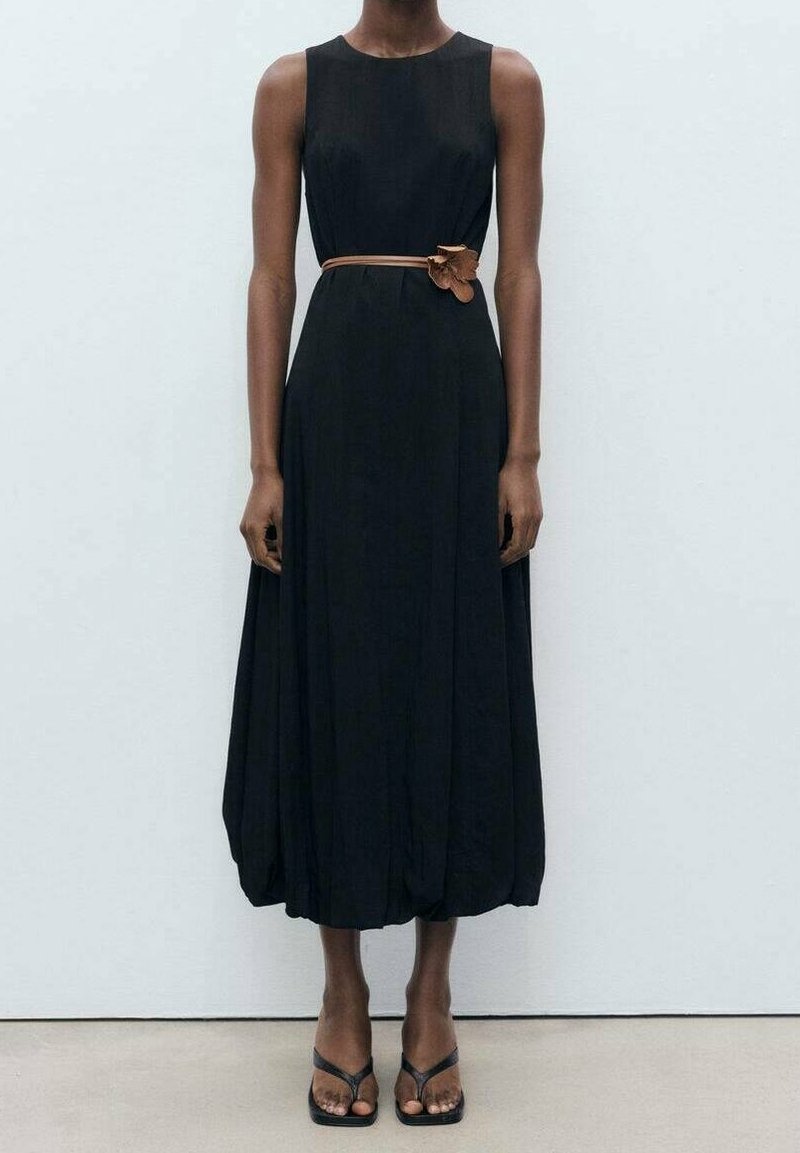 Personne portant une robe midi noire sans manches avec une ceinture marron et des sandales noires à bout carré, debout devant un fond blanc uni.