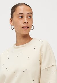 Μπεζ φούτερ με στρογγυλή λαιμόκοψη, διακοσμητικά τσιμέντα και μαλακή υφή. Ο σχεδιασμός περιλαμβάνει διάσπαρτες μεταλλικές λεπτομέρειες.