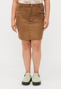 Brun semsket miniskjørt med en tettsittende design, frontlommer og knappelukking øverst, kombinert med lysegrønne slip-on sko.
