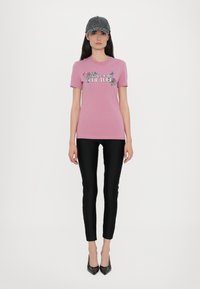 T-shirt di cotone rosa con grafica floreale e testo "Versace Jeans Couture". Indossato con leggings neri e tacchi neri a punta; berretto grigio.