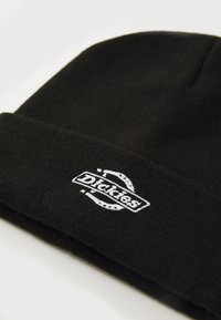 Melnā adītā cepure ar saliekamu maliņu, kurai ir balts izšūts "Dickies" logo un apaļie akcenti. Mīksta tekstūra un bezšuvju dizains.