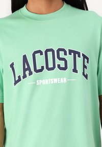 Mėtų žalia medvilninė marškinėliai trumpomis rankovėmis, su išryškintu „LACOSTE“ tamsiai mėlyna spalva ir „SPORTSWEAR“ baltu tekstu žemiau.