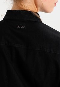 Camicia in denim nero con colletto classico, cucitura a contrasto sulla schiena e un logo discreto impreziosito da strass sulla parte superiore della schiena.